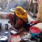 A Pakistani woman is shown preparing bread and rolling it out in a work station on the ground outside. She is surrounded by pots and pans and next to a small fire with a plate on top for cooking.