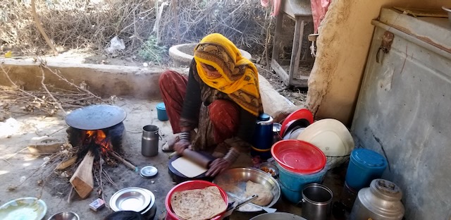 A Pakistani woman is shown preparing bread and rolling it out in a work station on the ground outside. She is surrounded by pots and pans and next to a small fire with a plate on top for cooking.