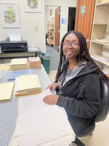 A young black student with long braids, black sweatshirt and glasses works in Loretto's archives. She is seen going through papers.