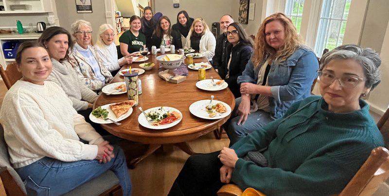 13 people smile for a picture before enjoying a family-style dinner together in dining room of a house.