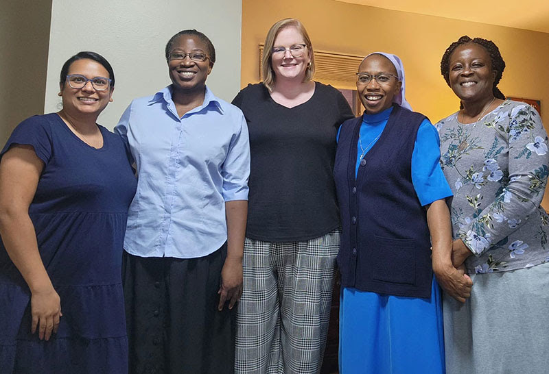 Five women stand in a row smiling at the camera together. There are three African women, one Pakistani woman and one white woman.