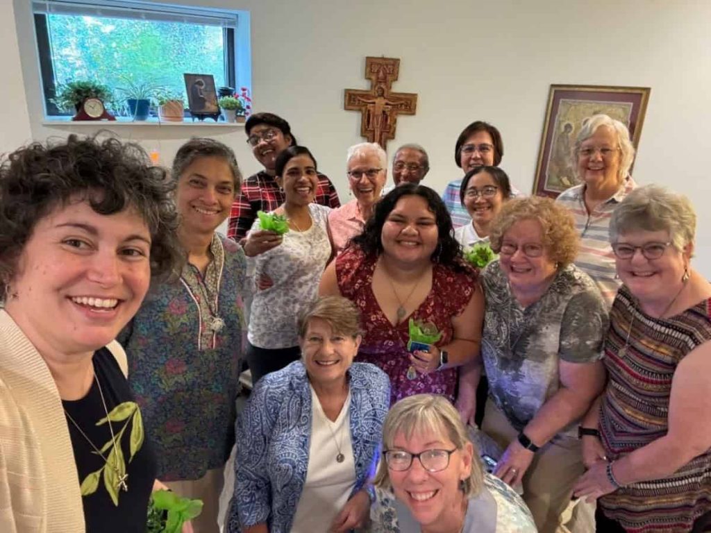 A group of women of various ages and races smile for a photo in front of a crucifix. They are holding small green plants.