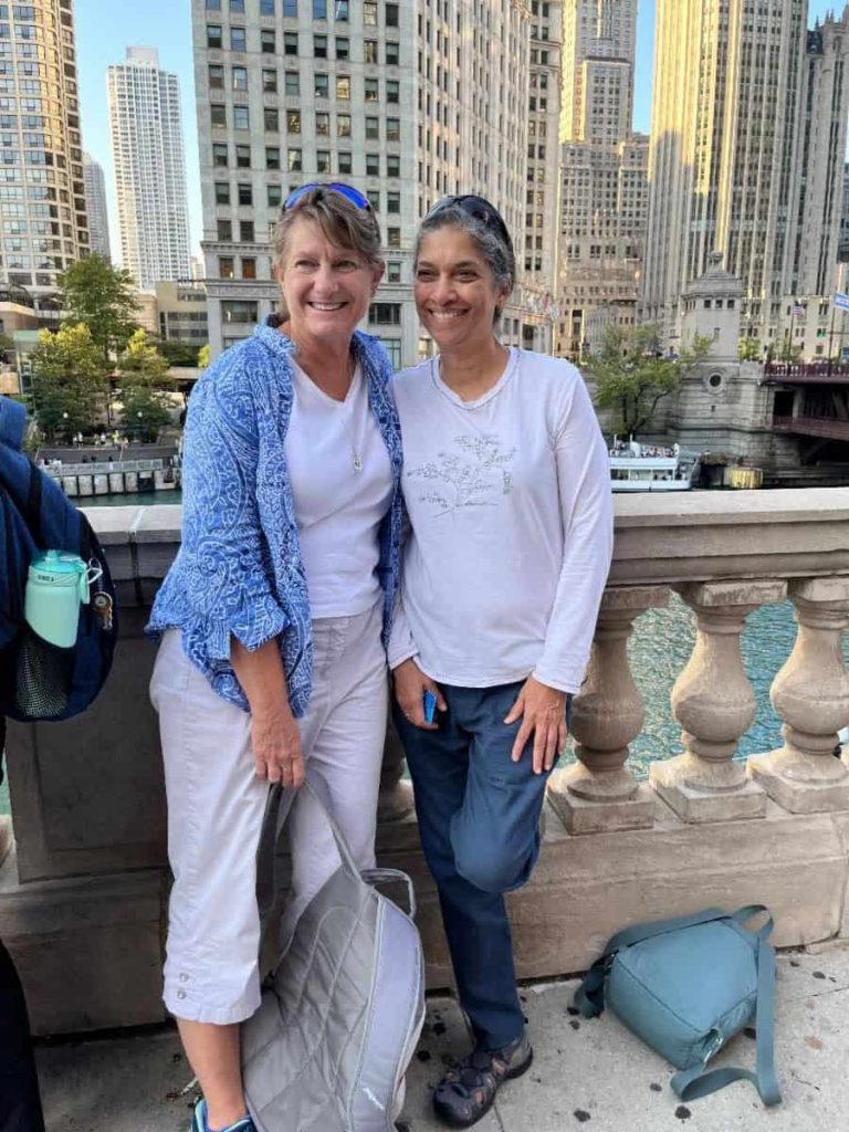 Two middle agesd women stand next to each other in casual clothing and sunglasses on their head smiling for a photo outside of downtown Chicago. You can see tall buildings behind them.