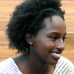 A young Black woman with medium length natural hair and two small silver earrings in her right ear is smiling looking to the side and wearing a light blue and white striped shirt.