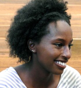 A young Black woman with medium length natural hair and two small silver earrings in her right ear is smiling looking to the side and wearing a light blue and white striped shirt.