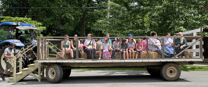 Many children in adults in brightly colored clothing are seen sitting on top of a hay wagon as they are about to take a tour of a farm on a sunny day.