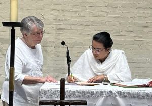 An older woman in white looks on as a younger woman with dark hair and traditional Pakistani clothing signs a document on an altar.