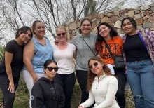 Eight women smile for a group photo on a casual hike on a cloudy day with six women standing in the back and two women squatting in the front wearing sunglasses.