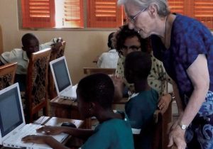 An older woman in a blue dress and glasses stands over a young student working on a computer to offer support.