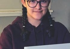 A high school student with big black round glasses and braids smiles softly while wearing a dark purple sweatshirt
