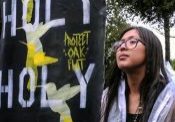 A young indigenous girl with big glasses, long dark hair and a necklace with a shell on it stands powerfully holding a sign that says "Holy Holy Holy Holy Protect Oak Flat."