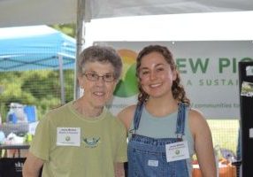 From left, an older woman with grey hair, glasses and a green t-shirt stands smiling with a young woman with brown curly hair wearing a blue tank top and jean overalls. They are standing together in front of a sign that says "new pioneers for a sustainable future."